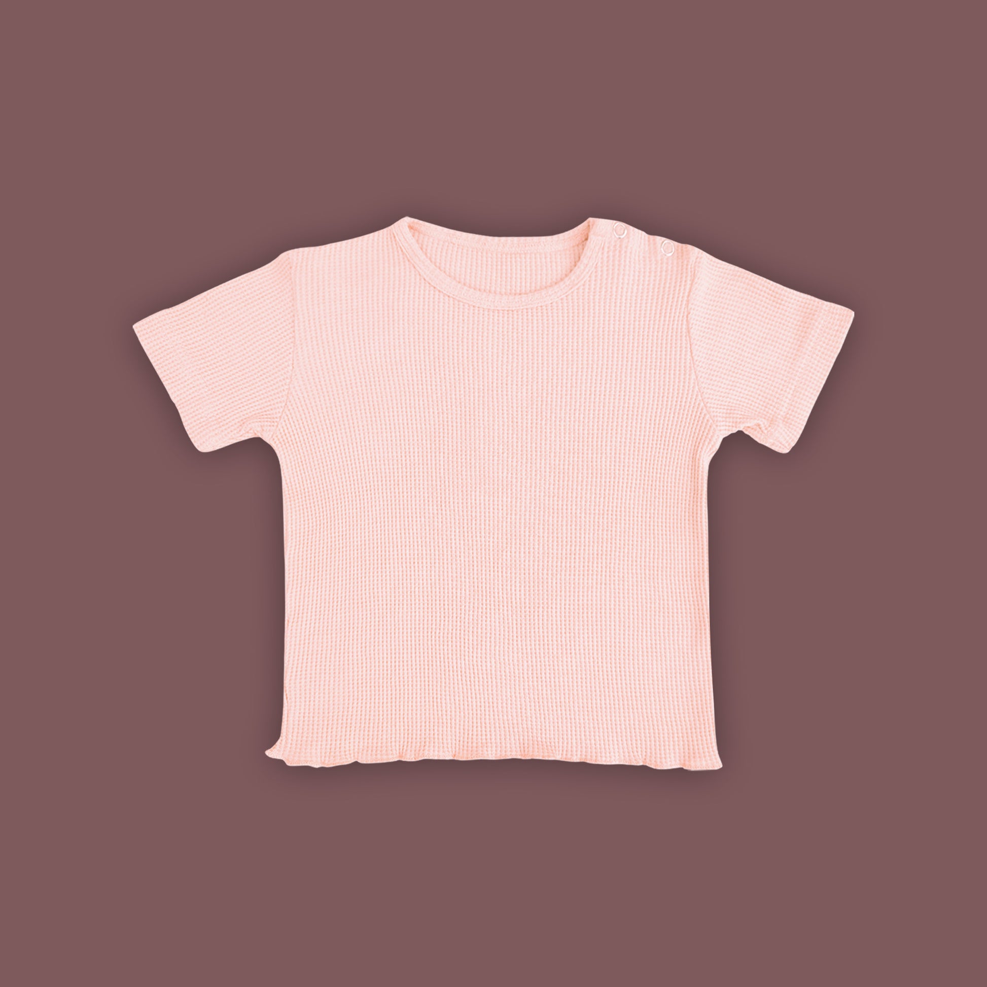 A baby short-sleeved T-shirt in a soft pastel pink color, displayed against a muted brown background. The T-shirt is made from ribbed fabric and features snap buttons on one shoulder for easy dressing. The hem has a subtle wavy finish, adding a delicate touch to the design.