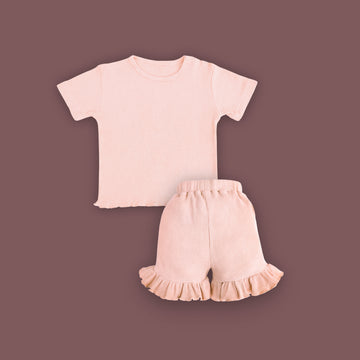 A baby clothing set consisting of a short-sleeved, ribbed T-shirt and ruffled shorts in a soft pastel pink color, displayed on a muted brown background. The T-shirt features snap buttons on one shoulder for easy dressing, and the shorts have an elastic waistband with ruffled hems for a cute and comfortable look.