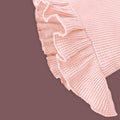 A close-up view of a baby garment in soft pastel pink, highlighting the delicate ruffled hem detail. The fabric has a ribbed texture, and the ruffles add a charming, stylish touch. The muted brown background provides contrast, emphasizing the intricate design and stitching.
