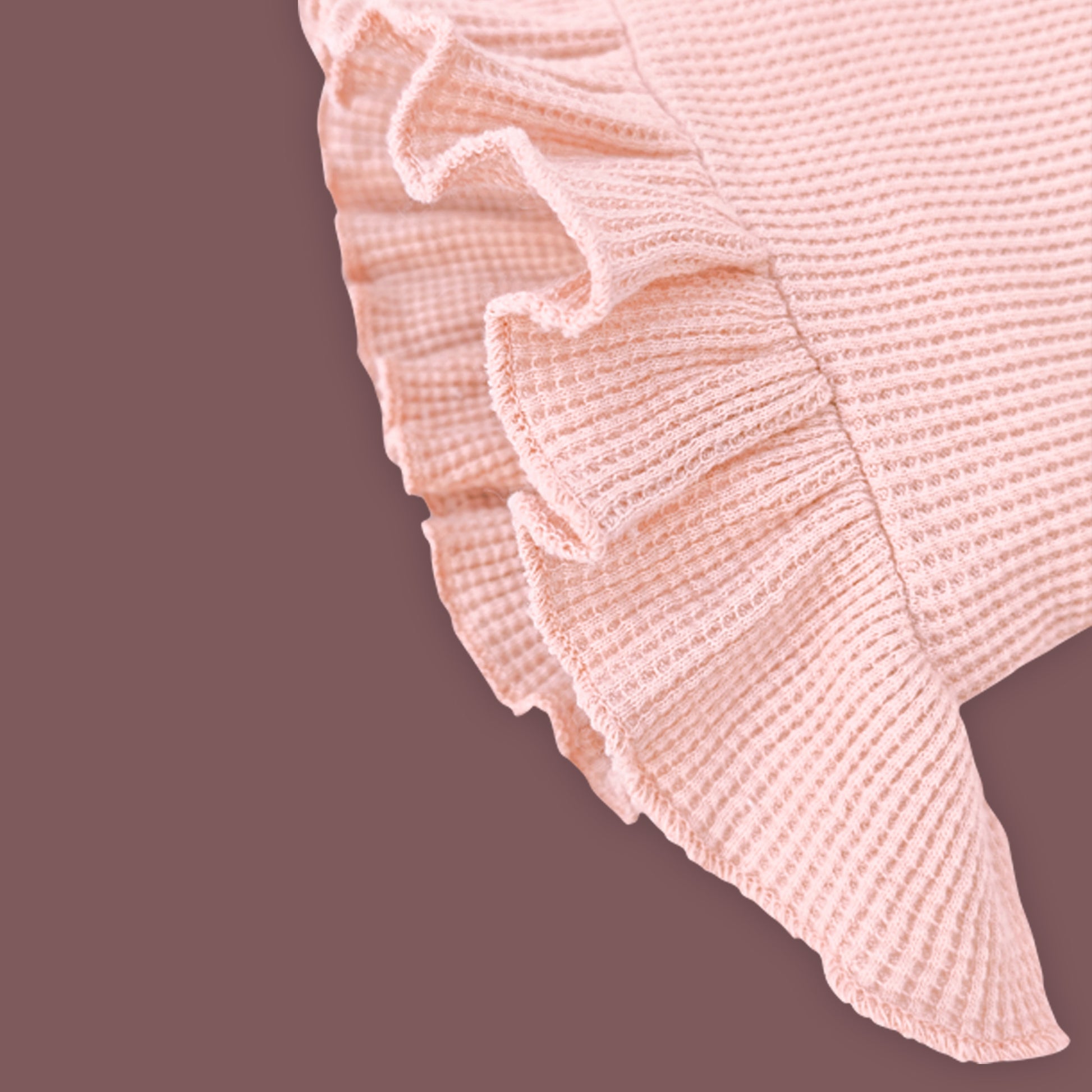 A close-up view of a baby garment in soft pastel pink, highlighting the delicate ruffled hem detail. The fabric has a ribbed texture, and the ruffles add a charming, stylish touch. The muted brown background provides contrast, emphasizing the intricate design and stitching.