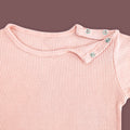 A close-up view of a baby T-shirt in soft pastel pink, highlighting the ribbed fabric texture and shoulder snap button closure. The two metallic snap buttons on one side allow for easy dressing and a comfortable fit. The delicate stitching and gentle stretch of the material are emphasized against a muted brown background.