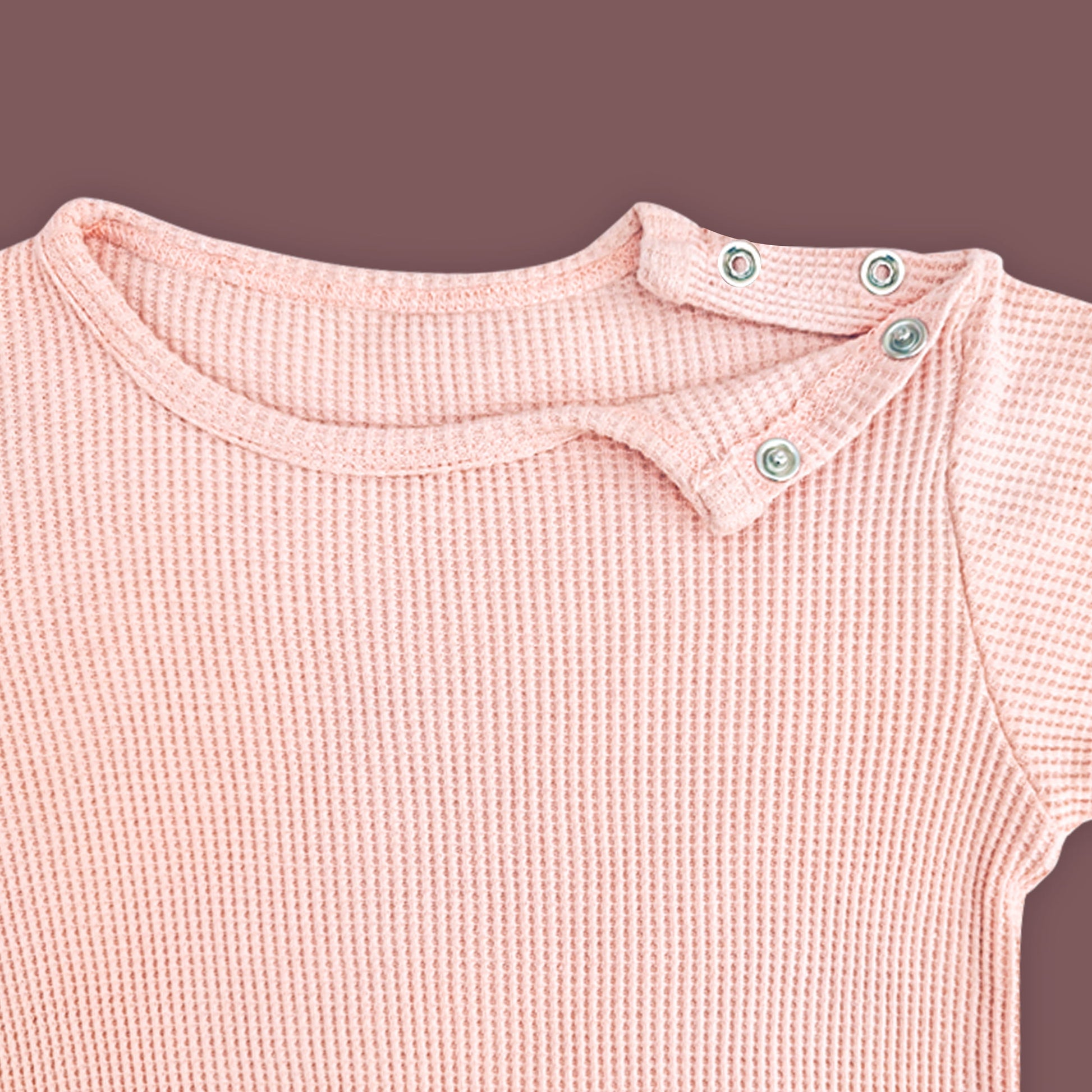 A close-up view of a baby T-shirt in soft pastel pink, highlighting the ribbed fabric texture and shoulder snap button closure. The two metallic snap buttons on one side allow for easy dressing and a comfortable fit. The delicate stitching and gentle stretch of the material are emphasized against a muted brown background.